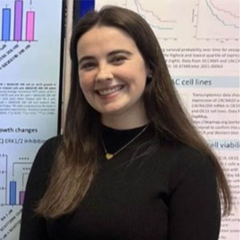 Ellie Power in a black mockneck top smiling at the camera in front of a wall of posters containing scientific information.